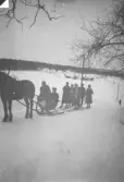 Ett sällskap färdas vintertid på en snötäckt landsväg med häst och släde. Några personer sitter i släden medan andra står bredvid på skidor. Öppet landskap med gårdsbebyggelse och telefonstolpar i bakgrunden.