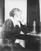 En kvinna sitter vid ett bord i en interiör och lutar hakan mot handen. På bordet står en karaff på en bricka. Mörka möbler och väggparti i bakgrunden.