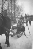 Ett sällskap färdas vintertid på en snötäckt väg med häst och släde. Ett barn sitter i släden medan en kvinna står bredvid på skidor och håller i en stav. Ytterligare personer syns längre bak på vägen. Träd och bebyggelse i bakgrunden.