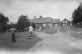 En gårdsmiljö med flera trähus. I förgrunden står två män, varav en arbetar med mätutrustning på marken. I bakgrunden syns personer vid husen samt en grusväg som leder fram till byggnaderna.