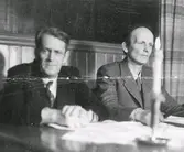 Två män som sitter nedanför en scen med ett levande ljus tänt på bordet framför dem. Mannen till vänster är folkskolläraren Anders Nordin (se bildnr FHN776) och de sitter troligen i Tvååkers Medborgarhus/Folkets Hus. Uppgifter saknas. Tillhör samlingen med fotokopior från Hallands Nyheter som är från 1930-1940-talen.