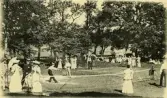 Tennisspel och umgänge kring ett tennisnät i parkmiljö på Särö. Några kvinnor har solparasoll.