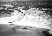 Flygbild över Åsa. Sandstranden med kiosken centralt placerad och campingen en bit upp från vattnet. Solande och badande människor. I bakgrunden breder ortsbebgyggelsen med villor och sommarstugor ut sig i terrängen.