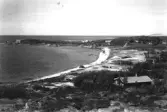 Åsa Skytteviken. Vy från bergen över havsviken med sandstrand och några båtar förtöjda i vattnet. Strödda i terrängen ligger fritidshus.