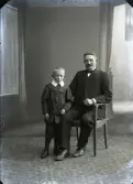 Familjebild, far och son i helfigur där fadern sitter. Ateljébild. Beställare: J A Johansson, Åsa.