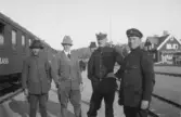 Fyra män stående vid järnvägsspår intill ett tåg. Bebyggelse syns i bakgrunden.