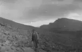 En person vandrar genom ett stenigt fjällandskap med bergsryggar i bakgrunden. Terrängen består av lösa stenar och branta sluttningar.