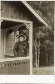 Operasångerskan Christina Nilsson i mörk sidendräkt och ljus hatt. Hon står på verandan till ett enkelt rödmålat hus med 
vita knutar.
Möjl. Loka brunn (?).