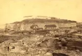 Stenbrottet nedanför Varbergs fästning. I förgrunden ses små skjul och några stenhuggare och barn. Troligen 1880-1890-tal.