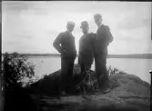Tre män och hund står på klippa vid vattnet, Uppland