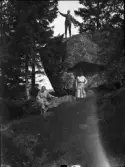 Man står högt upp på en sten i skogen, två kvinnor står nedanför, Uppland