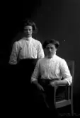 Gruppbild, två kvinnor i 3/4 figur där en sitter. Ateljébild. Beställare: Anny Bengtsson, Himle.