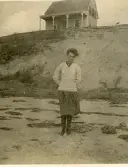 Porträtt av en kvinna, vid namn Lizzie, som står på en strand med en stuga i bakgrunden vid Wellfleet Cape cod, USA  (enligt en del av texten på baksida kort).
