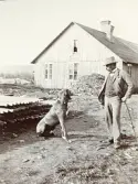 Man med kostym och en hund, står framför en arbetarbostad vid bygget av Älvkarleby kraftverk i Älvkarleby, omkring år 1906 (enligt text i album).