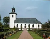Exteriörbild. Färgaryds kyrka och kyrkogård sedd från söder med grusgången som leder rakt mot den centrerade porten i långskeppet. Kyrkan uppfördes 1845 – 1846 i nyklassicistisk stil efter ritningar av byggmästare Petter Pettersson, som också ledde arbetet.