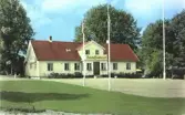 Kolorerad exteriörbild av Hishults gästgivaregård. Tre flaggstänger framför huset, med svenska och amerikanska flaggor. (Se även bildnr FFAD448.)