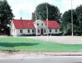 Exteriörbild av Hishults gästgivargård, vilket förkunnas på balkongräcket över entrén. I förgrundet står ett par flaggstänger med svenska och amerikanska flaggorna. (Se även bildnr FFAD449.)