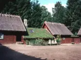 Exteriörbild av Gammelgården i Hishult. En kringbyggd 1700-talsgård som flyttats till Hishult på 1950-talet. Hembygdsgård. Rödmålad högloftsstuga, ryggåsstugan har grästak och de två loften halmtak. Framför ett av husen finns en plantering omgiven av ett staket samt bikupor. I bakgrunden granskog.