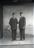 Ateljébild av två män i helfigur i uniform med skärmmössor med texten 