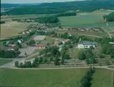 Flygbild över Slättåkra. Slättåkra kyrka och kyrkogård med omgivand bebyggelse, odlingslandskap och skogsklädda åsar.
