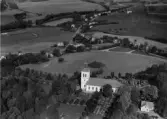 Flygbild över Vessige kyrka och kyrkogård. Bostadshus, skog och odlingslandskap. Huset till höger om kyrkan är troligen Klockaregården. Landsvägskorsning i centrum av bilden där Möllegårdsvägen möter Sjönevadsvägen. Vid korsningen ligger Loricks smedja och idrottsplatsen Brovallen ses i vänster kant. Ån Ätran i bakgrunden.