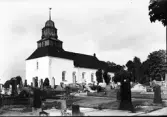 Exteriörbild av Slättåkra kyrka och kyrkogård. Kyrkan har tidigmedeltida ursprung. Det låga tornet uppfördes 1776 i samma bredd som långhuset. Den spånklädda lanterninen har upptill en åttakantig form med ansvällning nedtill och klockformad huv.