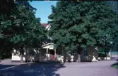 Exteriörbild av Sjönavads gästgivaregård omgiven av träd tätt inpå byggnaden. Entré med farstukvist.