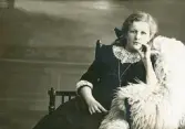 En flicka i mörk klänning och spetskrage sitter i en s.k. munkstol hos fotografen. 
Trol. Ruth Svensson (1901-1991), Alvesta, senare Stockholm.
