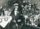 En student i smoking och studentmössa, avfotograferad i ett vardagsrum, omgiven av blommor.
Seved August Svensson (1894-1962), senare postassistent och kamrer, Skurup.