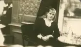 En tonårsflicka i mörk klänning sitter vid ett caféfbord.
