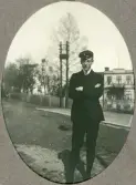 En ung man i mörk kavaj, knäbyxor och skolmössa poserar vid en gata (möjl. husen Ericsberg och Fridsberg vid Kungsvägen, Växjö).