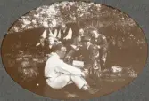 Några herrar i kostym och hatt m.m. har slagit sig ner i en skogsbacke en sommardag. 
En biologiexkursion för folkskollärare (?).