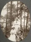 Två kvinnor i ljusa sommarklänningar och en man i kostym har slagit sig ner på en sten i skogen. I bakgrunden skymtar en sjö. 
Möjl. fru Helga Svensson med dotter Ruth och en okänd man.