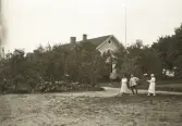 Tre personer framför gaveln till Östra folkskolan i Alvesta, folkskollärare Amos Svenssons hem under många år. I bakgrunden skymtar Aringsås (Alvesta) kyrka.
Längst till höger Ruth Svensson.