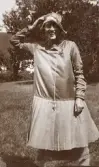 En glad ung kvinna poserar i en trädgård. I bakgrunden skymtar Aringsås (Alvesta) kyrka.
Trol. Ruth eller (möjl.) Blenda Svensson.