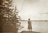En ung kvinna i kappa och hatt (Ruth Svensson?) poserar på en bergsknalle vid en sjö.