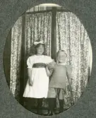 Två små barn, en flicka och en pojke, poserar framför en skärm med småblommigt tyg. 
Något suddigt foto.