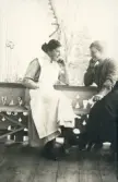 En sjuksköterska och en annan ung kvinna i blus och kjol sitter på en balkong och pratar.