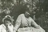 Två unga kvinnor i rutiga och randiga sommarklänningar m.m. tittar tankfullt mot fotografen.
Kvinnan till vänster - Ruth Svensson.