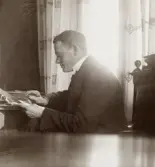 En man i kostym och stärkkrage sitter och studerar en skrivelse vid ett skrivbord.