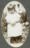 En liten flicka i ljus sommarklänning poserar i en trädgård. I bakgrunden syns två pojkar (bröder?) och en liten flicka.