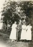 Tre kvinnor i ljusa sommarklänningar och en (och en halv) man i kostym och hatt poserar vid en gärdesgård.