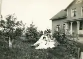 Fyra kvinnor i ljusa sommarklänningar och två män i kostym m.m. har slagit sig ner på en gräsmatta i en trädgård. I bakgrunden syns ett bostadshus med veranda.
