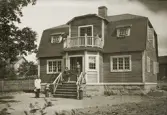 Ett nybyggt bostadshus med trädgård. På trappan framför huset syns en medelålders man och en kvinna. Till vänster kommer en man via gårdsplanen med leverans av en tidning m.m.
Kalmar (?).