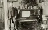 En man i kostym med stärkkrage och ljus väst poserar vid ett piano. 
Pianot verkar vara levererat av firman 