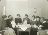 En syförening (?). Sju kvinnor med diverse handarbeten har samlats kring ett bord.