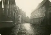 En regnvåt gata i Ystad (?) med korsvirkeshus m.m. På en skylt till höger kan man läsa: 