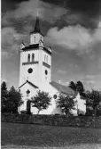 Exteriörbild av Knäreds kyrka och kyrkogård, omgiven av träd och stenmur. Urtavla på kyrktornet. Kyrkan ritades av Hans Jakob Strömberg och uppfördes 1853 i vitputsad gråsten av byggmästare Anders Johansson från Bredareds socken. Invigningen ägde rum på bönsöndagen den 8 oktober 1854. Förlag: J. Alfr. Jacobsson, Knäred