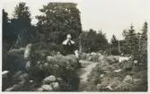 En kvinna i hatt och kappa med stor krage sitter vid ett stenparti i en park (botanisk trädgård?).