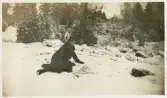 En kvinna i rutig kappa och hatt poserar i en snöig slänt, med en gärdesgård i bakgrunden.